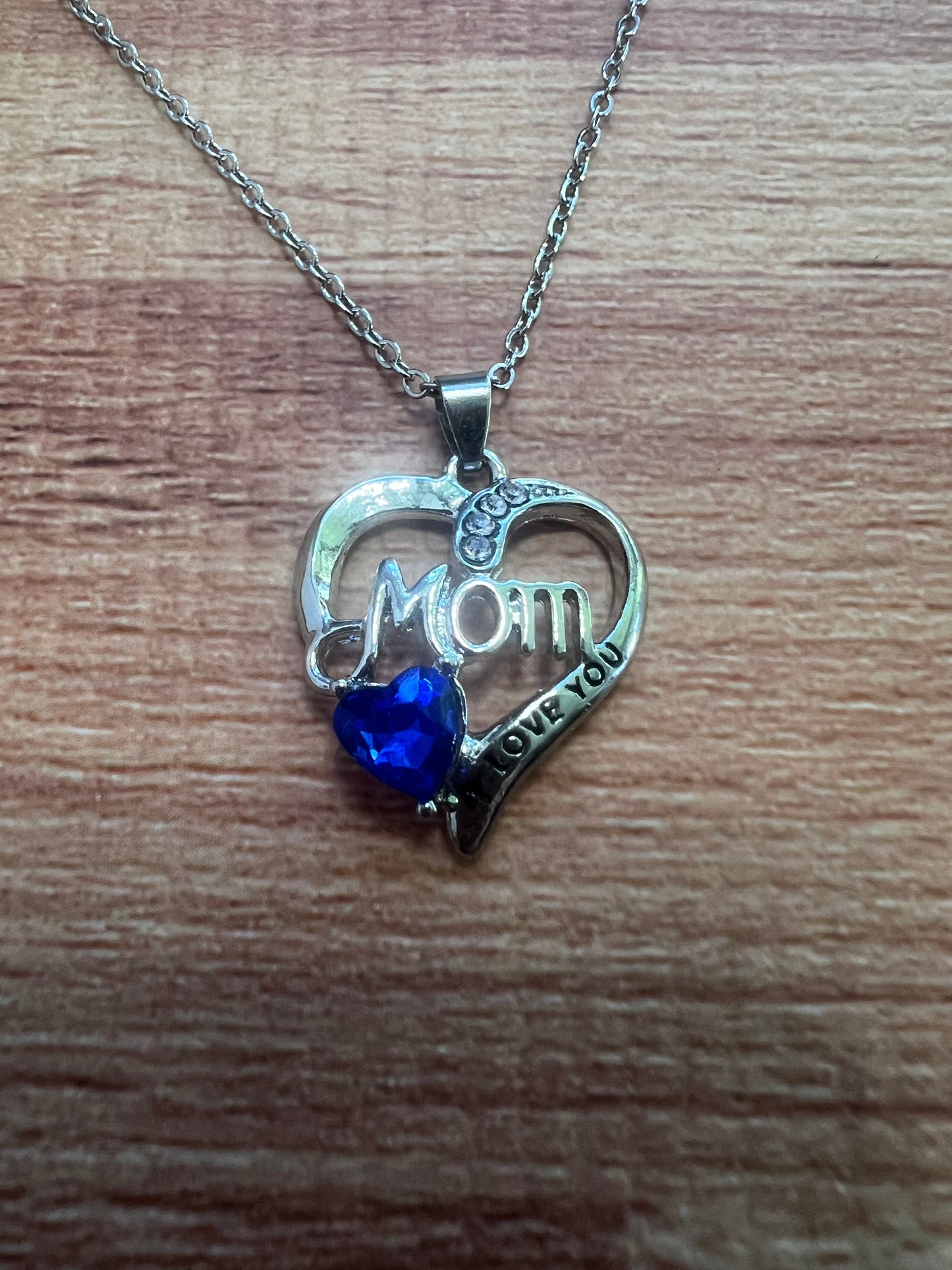 Heart-shaped 'Mom' pendant with blue gem on a chain against a wooden background