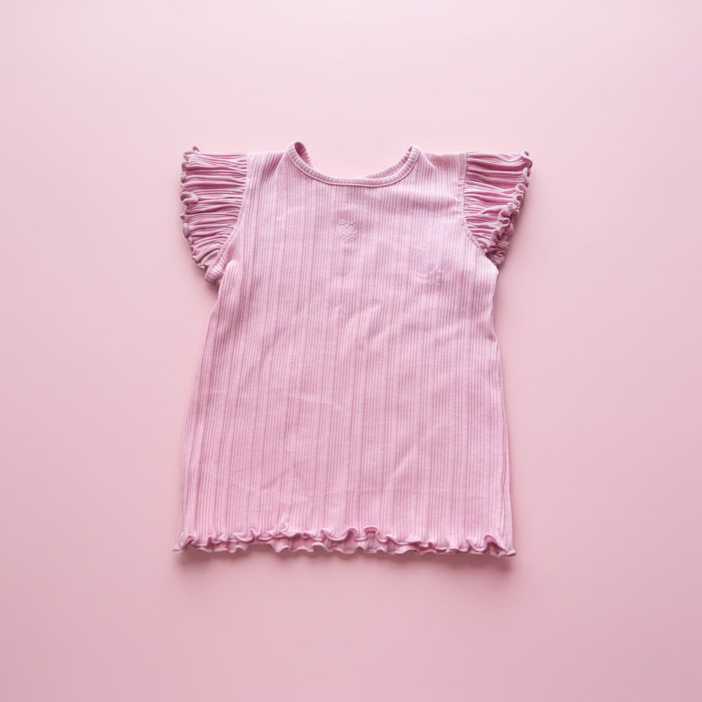 Pink ruffled top on a pink surface with beige pants and cushion.